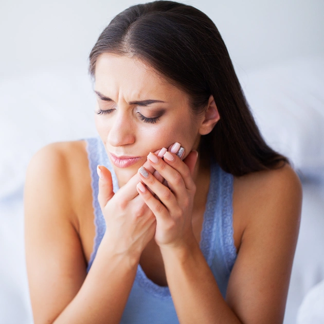 Woman holding cheek in pain, appearing to experience dental discomfort indoors.
