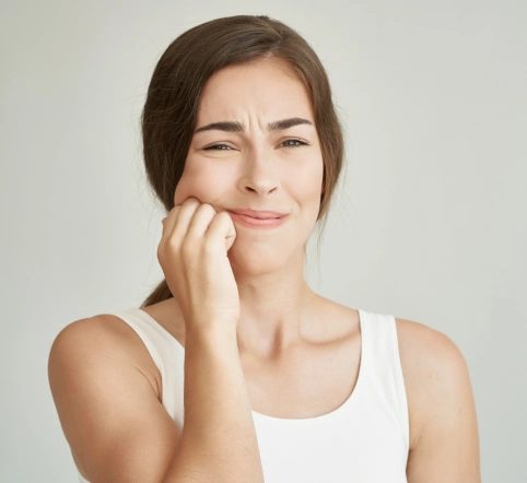 Woman in white tank top with hand to face, appearing thoughtful against neutral background.