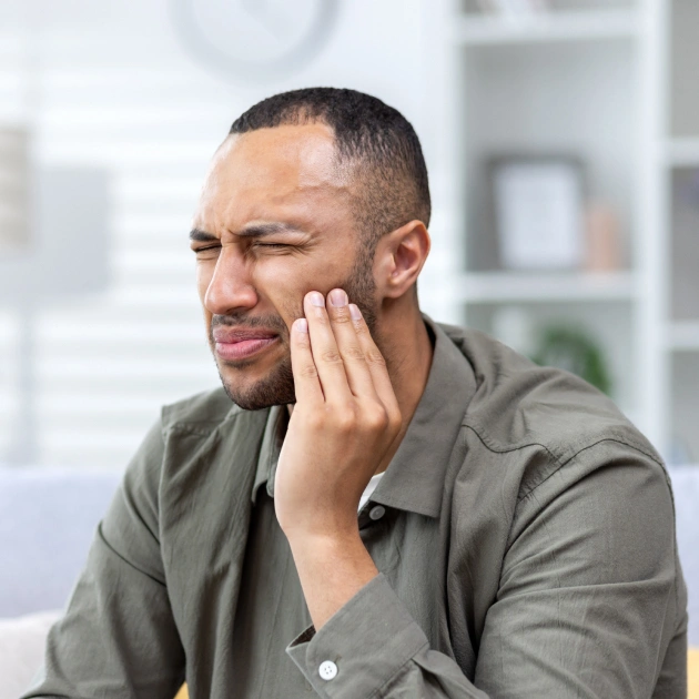 Man in gray shirt touching jaw in dental office, appearing to experience tooth pain or discomfort.